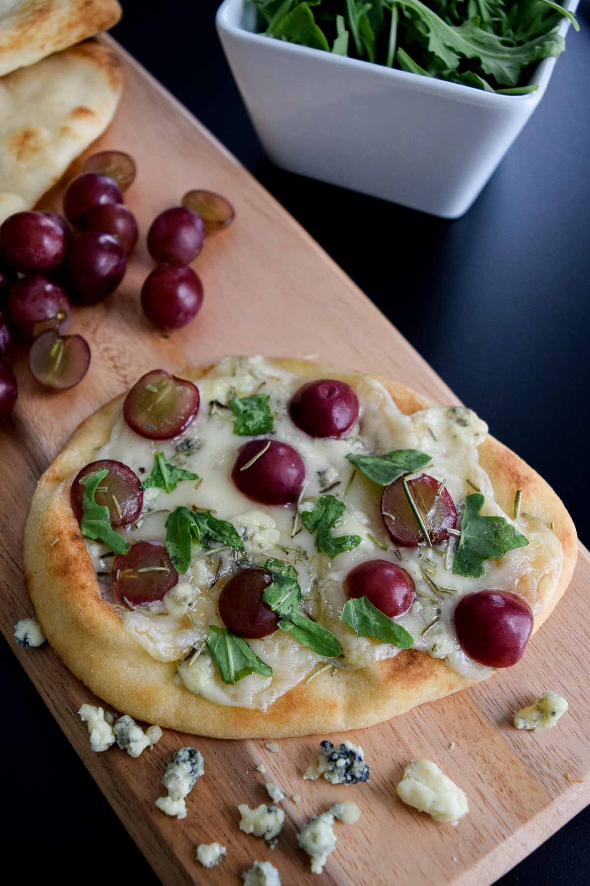 close up view of a flatbread topped with cheese grapes and arugula sitting on a wooden serving board